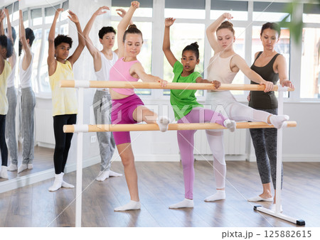 Teenage girls doing leg stretching exercise holding by ballet barre during dance courses 125882615