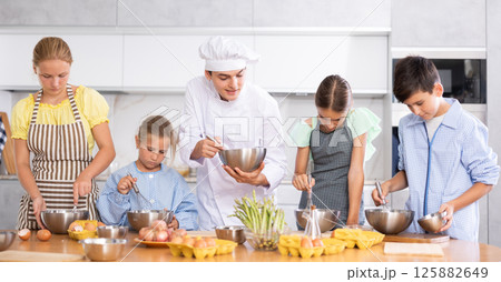 Young guy cook teaches to cook group of children Young guy cook teaches to cook group of children 125882649