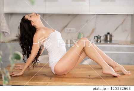 Portrait of happy brunette young woman wearing underwear playfully posing on table at white kitchen 125882668