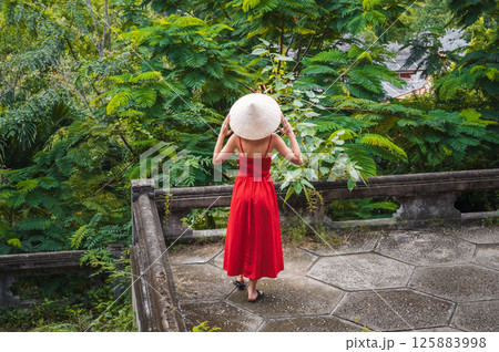 back of girl tourist traveler in a Vietnamese non la hat in red dress in garden. Tourism and travel in Vietnam 125883998