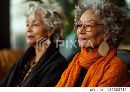 Two elderly women with gray curly hair, one wearing glasses and an orange scarf, sit indoors looking forward. Concept of wisdom and friendship. For senior lifestyle photo. 125888715
