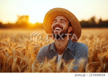 Man in straw hat laughing joyfully in golden wheat field during sunset. Concept of happiness and nature connection. For promoting rural lifestyle. 125889026