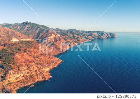 Rocky seashore with clear blue sky. Seascape in the evening. Aerial view from Maro Beach to Pigeon Cave and Cerro Gordo viewpoint. La Miel Viaduct on Mediterraneo Highway. Malaga, Granada, Spain 125890573