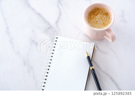 コーヒーを飲みながら、リング式のメモ帳とペンでデスクワークをしているイメージ 125892385