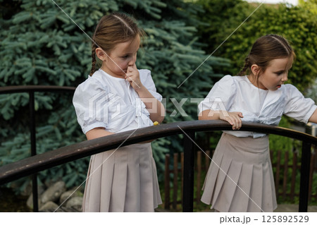 The twins observe the snail moving along the railing. Attentive girls in identical outfits create a sense of harmony. The twins observe the snail moving along the railing. Attentive girls in identical outfits create a sense of harmony. 125892529