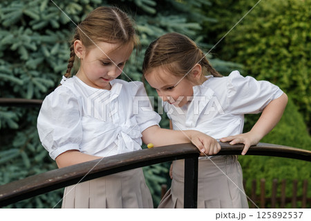 Little twins in light clothing lean over the snail. The girls stand near the railing, observing nature. Little twins in light clothing lean over the snail. The girls stand near the railing, observing nature. 125892537