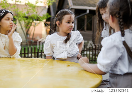 Cute girls are sitting at a children's table. The twin girls are dressed in white blouses, and their older sister is sitting next to them in a delicate dress. 125892541