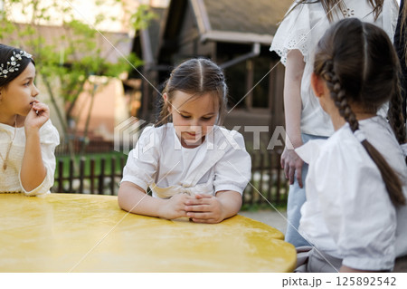 Three girls create a harmonious childhood scene at the bright table. The twin girls with braids sit attentively next to their older sister in white attire. 125892542