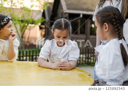 Three girls create a harmonious childhood scene at the bright table. The twin girls with braids sit attentively next to their older sister in white attire. 125892543