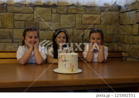 Three girls create a warm atmosphere near the brightly decorated cake. The twin girls with matching hairstyles sit close together, admiring the decorations. 125892551