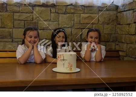 Three girls create a warm atmosphere near the brightly decorated cake. The twin girls with matching hairstyles sit close together, admiring the decorations. Three girls create a warm atmosphere near the brightly decorated cake. The twin girls with matching hairstyles sit close together, admiring the decorations. 125892552