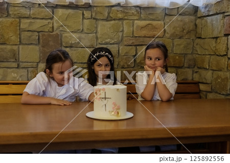 The girls in white clothing sit at the table near a delicious-looking cake. The twin girls with braids look touching beside the older girl. The girls in white clothing sit at the table near a delicious-looking cake. The twin girls with braids look touching beside the older girl. 125892556
