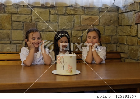 The girls in white clothing sit at the table near a delicious-looking cake. The twin girls with braids look touching beside the older girl. The girls in white clothing sit at the table near a delicious-looking cake. The twin girls with braids look touching beside the older girl. 125892557