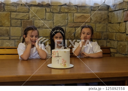 The girls in white clothing sit at the table near a delicious-looking cake. The twin girls with braids look touching beside the older girl. 125892558