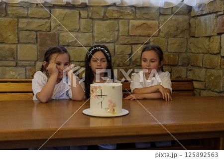 The twin girls with braids and the girl in a dress sit near the festive dessert. The girls appear focused and enchanted. The twin girls with braids and the girl in a dress sit near the festive dessert. The girls appear focused and enchanted. 125892563