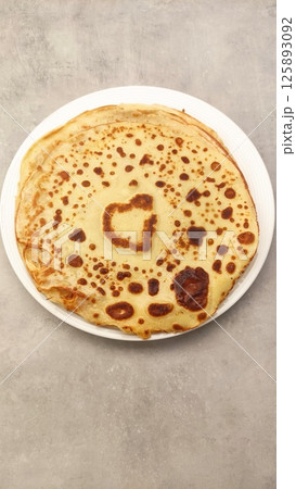 Golden-brown pancake with unique heart-shaped pattern on surface, served on white plate against textured gray background, perfect for breakfast or brunch culinary delight 125893092