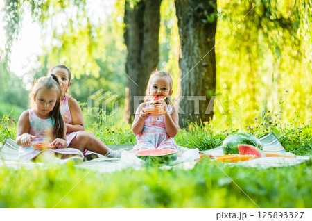 A funny portrait of incredibly small girls eating watermelon, a healthy fruit snack, a charming baby playing in a sunny garden on a warm summer day. A funny portrait of incredibly small girls eating watermelon, a healthy fruit snack, a charming baby playing in a sunny garden on a warm summer day. 125893327