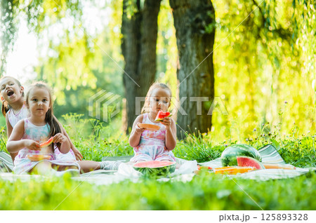 A funny portrait of incredibly small girls eating watermelon, a healthy fruit snack, a charming baby playing in a sunny garden on a warm summer day. A funny portrait of incredibly small girls eating watermelon, a healthy fruit snack, a charming baby playing in a sunny garden on a warm summer day. 125893328