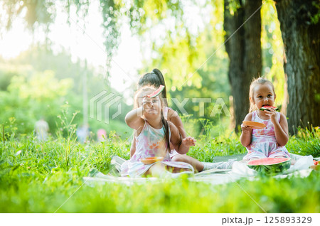 Three sisters are sitting on a soft blanket on the grass enjoying a sunny picnic. The girls are wearing matching dresses and sharing a juicy watermelon.  125893329
