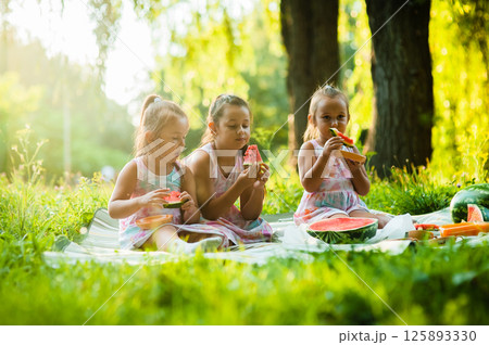 Three sisters are sitting on a soft blanket on the grass enjoying a sunny picnic. The girls are wearing matching dresses and sharing a juicy watermelon. Three sisters are sitting on a soft blanket on the grass enjoying a sunny picnic. The girls are wearing matching dresses and sharing a juicy watermelon. 125893330