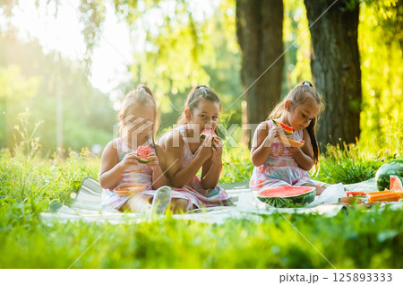 The girls are happily eating slices of watermelon while sitting in the shade. Their identical dresses make them look even more like best friends. The girls are happily eating slices of watermelon while sitting in the shade. Their identical dresses make them look even more like best friends. 125893333