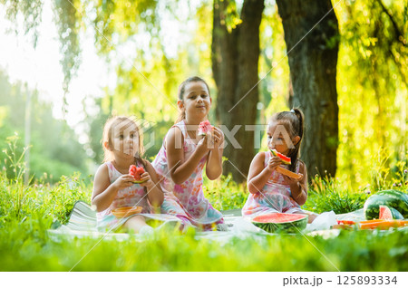 The girls are happily eating slices of watermelon while sitting in the shade. Their identical dresses make them look even more like best friends. The girls are happily eating slices of watermelon while sitting in the shade. Their identical dresses make them look even more like best friends. 125893334