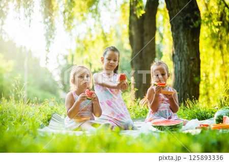 The picnic is filled with laughter as the sisters enjoy a watermelon. Their matching dresses make them look adorable. The picnic is filled with laughter as the sisters enjoy a watermelon. Their matching dresses make them look adorable. 125893336