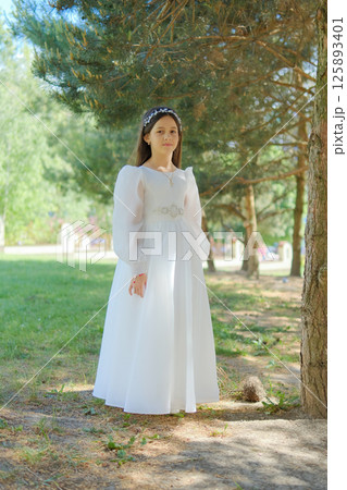 A little girl in an elegant dress stands among tall fir trees. The image of the girl combines tenderness, lightness and balance.  125893401