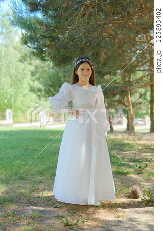 A girl in a formal dress is standing on a lawn under the sunlight. The white fabric of the dress shimmers against the green background.  125893402