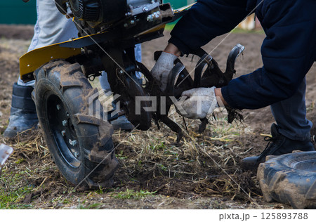 Preparing the land for planting with a walk-behind tractor. A farmer installs blades on a walk-behind tractor, preparing to plow well-weeded soil, preparing the land for the upcoming planting season. 125893788