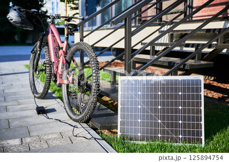 Pink electric mountain bike parked beside staircase, connected to small solar panel for charging. Bike rugged tires, mounted helmet on handlebar, situated on paved pathway with grassy lawn. 125894754