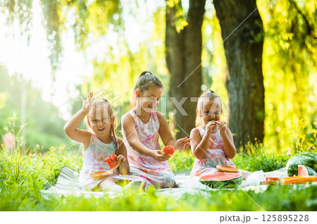 The sisters are sharing a picnic and laughing while eating watermelon. Their dresses match perfectly, making them look even more alike. The sisters are sharing a picnic and laughing while eating watermelon. Their dresses match perfectly, making them look even more alike. 125895228