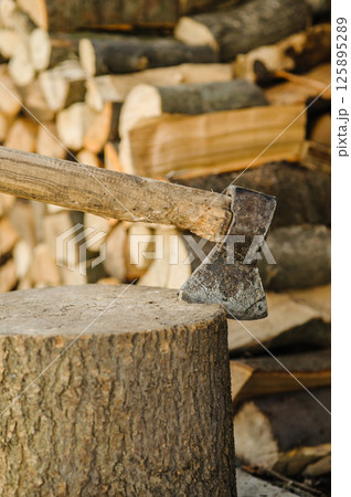 An axe is stuck in a sturdy stump. Behind it, there are many chopped and stacked logs. An axe is stuck in a sturdy stump. Behind it, there are many chopped and stacked logs. 125895289