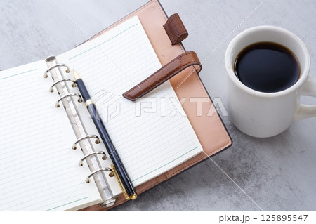 淹れたてのコーヒーを飲みながら、リング式のシステム手帳を開いてデスクワークをしているイメージ 淹れたてのコーヒーを飲みながら、リング式のシステム手帳を開いてデスクワークをしているイメージ 125895547