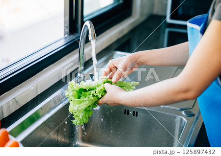 In a modern kitchen a woman's hands wash fresh vegetables under running water at the sink for preparing a vegan salad. Homemade healthy food concept with clean bright leafy greens. In a modern kitchen a woman's hands wash fresh vegetables under running water at the sink for preparing a vegan salad. Homemade healthy food concept with clean bright leafy greens. 125897432