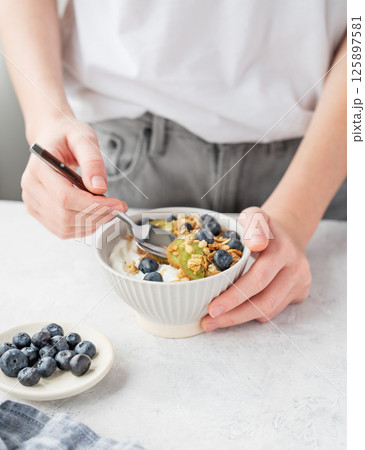 Bowl of granola with greek yogurt, kiwi and blueberries and spoon in hands of woman on white table. Bowl of granola with greek yogurt, kiwi and blueberries and spoon in hands of woman on white table. 125897581