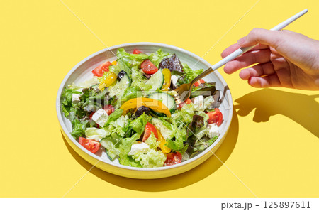 Hand holding fork vegetable salad with lettuce, olives and feta cheese on yellow background. 125897611
