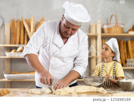Baker man cuts raw dough with knife. Little daughter helps him by sprinkling flour over dough through sieve 125898189