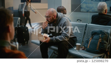 Crowded Airport Terminal: Man Eating Burger, Waiting for Delayed Airplane Flight, Going on Trip Crowded Airport Terminal: Man Eating Burger, Waiting for Delayed Airplane Flight, Going on Trip 125898270