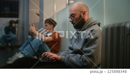 Man Working Online on Laptop, Sending Emails, Sitting on the Floor in Departure Lounge of Airline Hub Man Working Online on Laptop, Sending Emails, Sitting on the Floor in Departure Lounge of Airline Hub 125898348