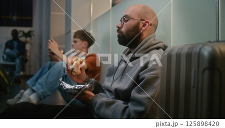 Man Eating Fast Food Burger while Sitting on the Floor in Departure Lounge of Airline Hub 125898420