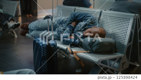 Tired Man with Suitcase and Backpack Sleeping on the Seats in Departure Lounge of Airport Terminal Tired Man with Suitcase and Backpack Sleeping on the Seats in Departure Lounge of Airport Terminal 125898424