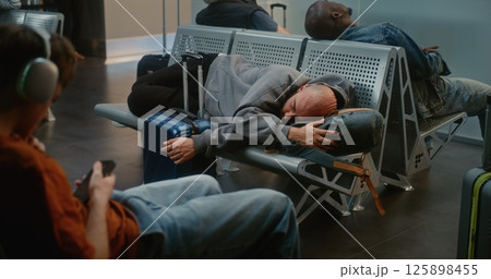 Crowded International Airport Terminal: Tired Man with Suitcase and Backpack Sleeping on the Seats 125898455