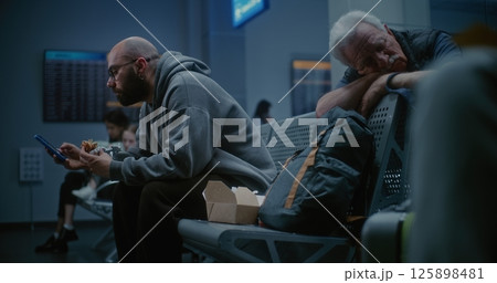 Boarding Lounge of Airport Terminal at Night: Man Eating Fast Food and Using Smartphone Boarding Lounge of Airport Terminal at Night: Man Eating Fast Food and Using Smartphone 125898481