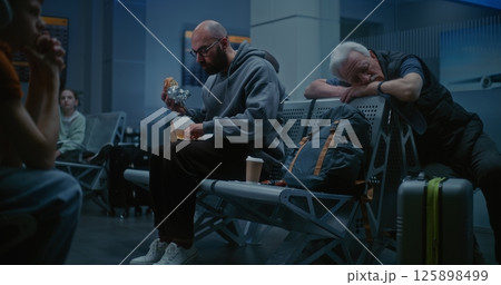 Man Eating Burger and Drinking Coffee Sitting in Departure Lounge of Airline Hub at Night Man Eating Burger and Drinking Coffee Sitting in Departure Lounge of Airline Hub at Night 125898499