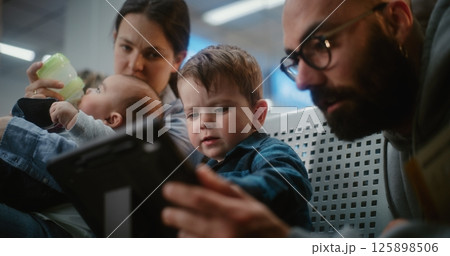 Airport Terminal: Woman Feeding Baby, Man Watching Cartoons on Digital Tablet with Little Son 125898506