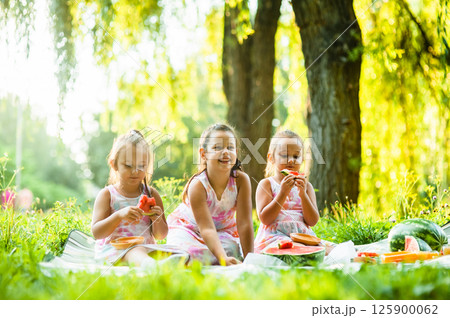 The three sisters are having a picnic surrounded by green grass and bright sunshine. The little twins share playful glances as they eat their watermelon, their dresses perfectly matching. The three sisters are having a picnic surrounded by green grass and bright sunshine. The little twins share playful glances as they eat their watermelon, their dresses perfectly matching. 125900062