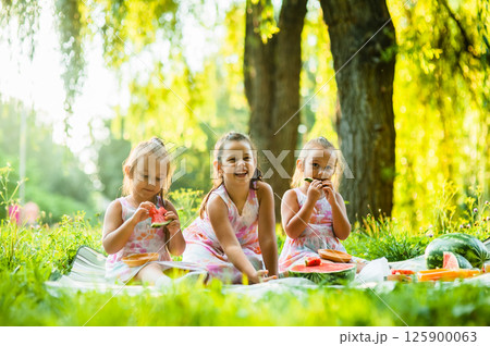 The sisters are sitting on a picnic blanket, enjoying a beautiful summer day. The twins giggle as they take big bites of watermelon, their matching dresses making them look even cuter. The sisters are sitting on a picnic blanket, enjoying a beautiful summer day. The twins giggle as they take big bites of watermelon, their matching dresses making them look even cuter. 125900063