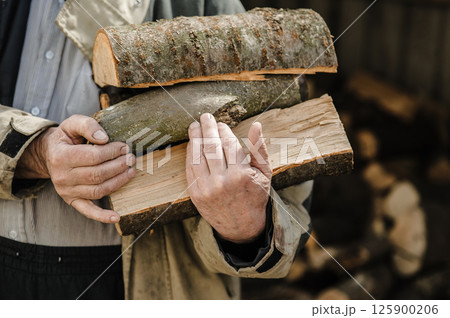 Rough, veined hands carry freshly chopped logs. Behind them, firewood is piled high. 125900206