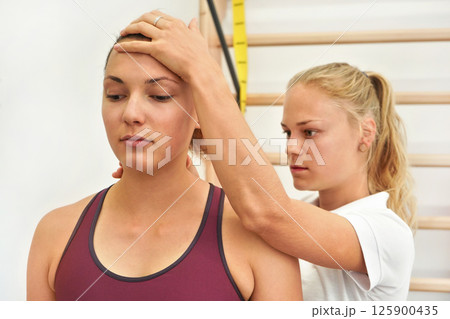 Young physiotherapist exercising with her female patient, fixing head and neck using hand, during exercise 125900435